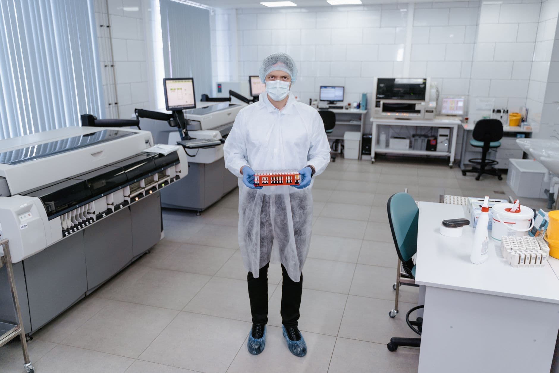Technician in full personal protective equipment carrying a sample rack through a clean laboratory